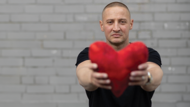 A man in a black t-shirt holds a stuffed heart towards the camera, with a brick wall in the background